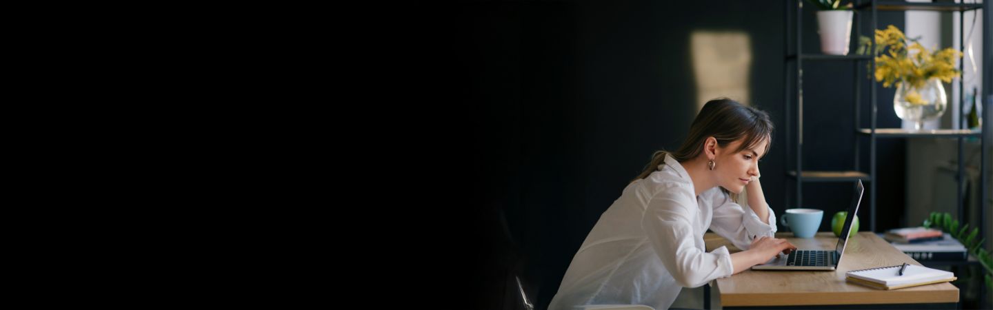 O tânără stă la un birou, lucrând concentrată la un laptop, cu un caiet și o ceașcă lângă ea. Fundalul este întunecat, iar accentul este pe tânăra femeie.