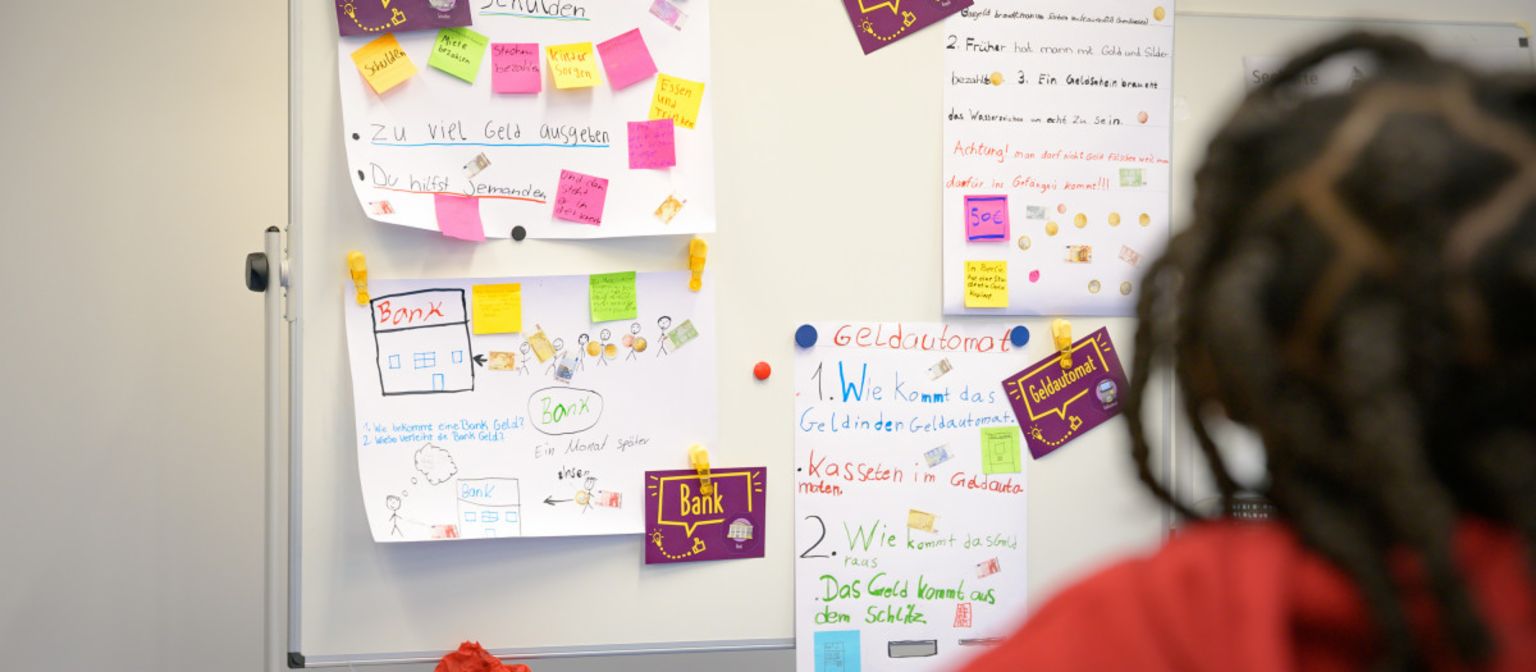 A photo shows post-its and paper notes on a board with terms like “Debt”, “Cash”, “Bank” and “ATM”. Underneath, the text reads: “In the ManoMoneta program developed by the EOS finlit foundation, schoolchildren aged 9 to 13 can learn about financial products.“