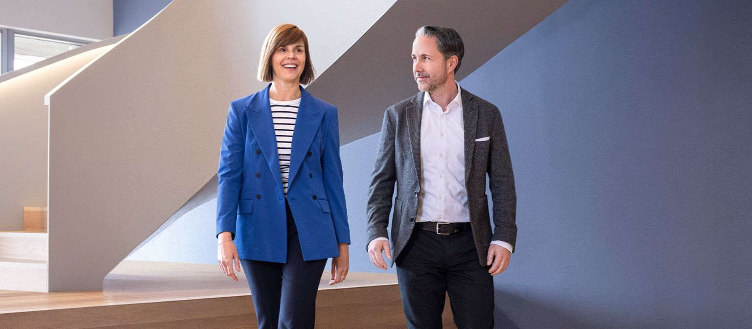 CEO-ul în blazer gri și CFO-ul Grupului EOS în blazer albastru trec printr-un birou modern. Spațiul dispune de feronerie mari, un perete curbat și lămpi suspendate elegante.
