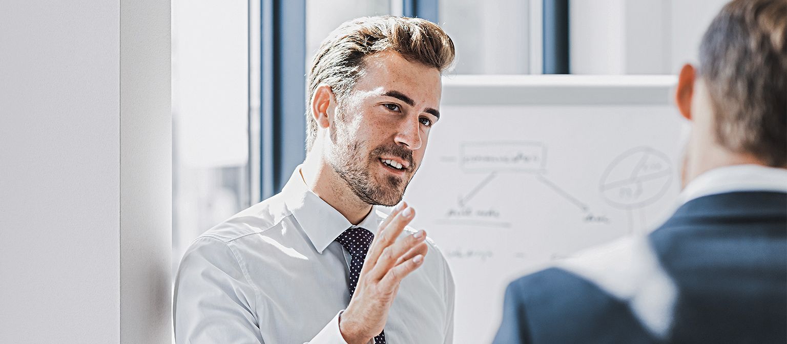Se vede un bărbat în ținută business care explică ceva cu o expresie prietenoasă și implicată. În fața lui se află o altă persoană care îl ascultă cu atenție. În fundal se vede o tablă albă cu o schiță.