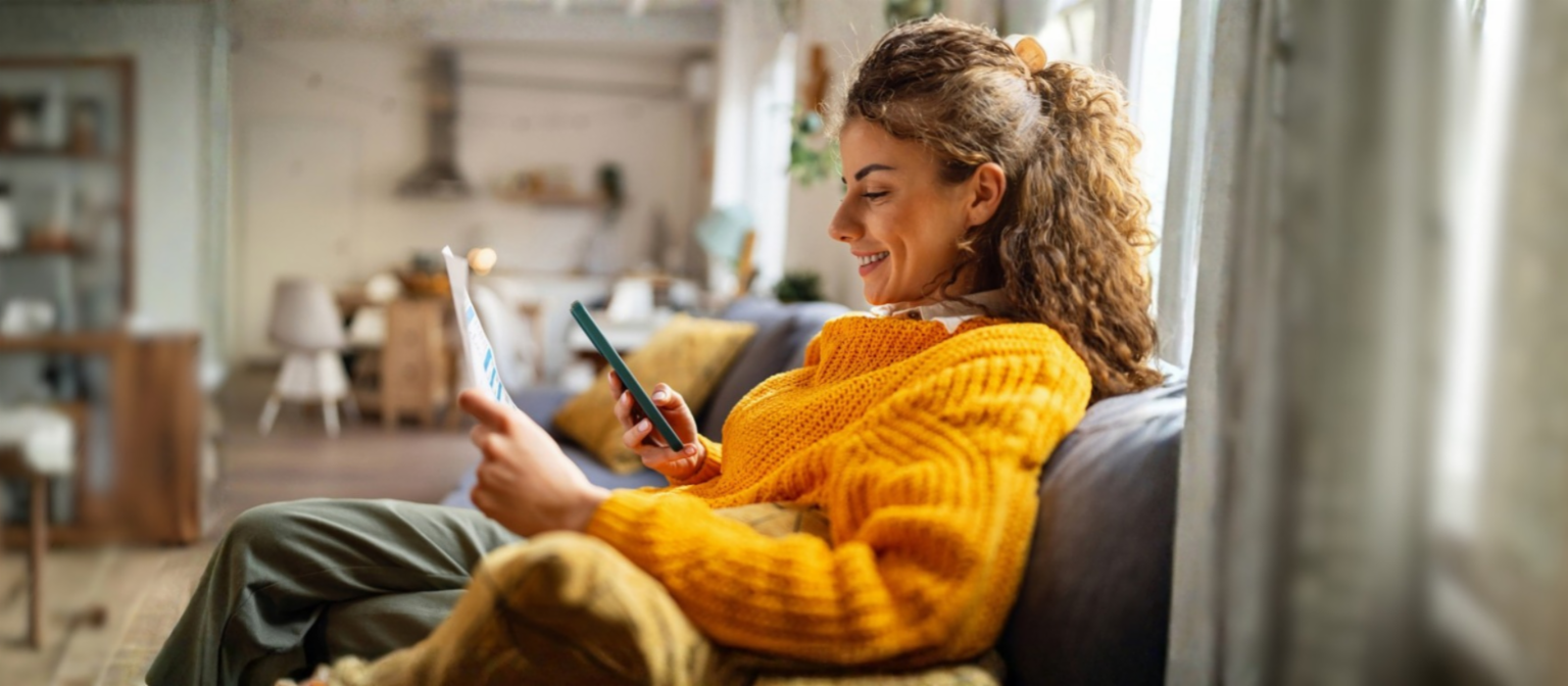 O tânără zâmbitoare cu păr creț, îmbrăcată într-un pulover galben confortabil, stă pe o canapea în sufrageria ei și se uită la smartphone-ul ei în timp ce ține un document de hârtie.