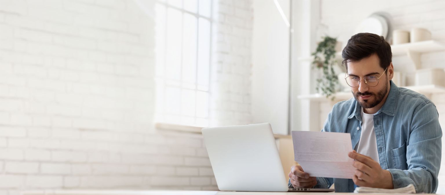 Bărbat cu ochelari lucrând la laptop în timp ce examinează documentul de reglementare a datoriilor într-o casă luminoasă