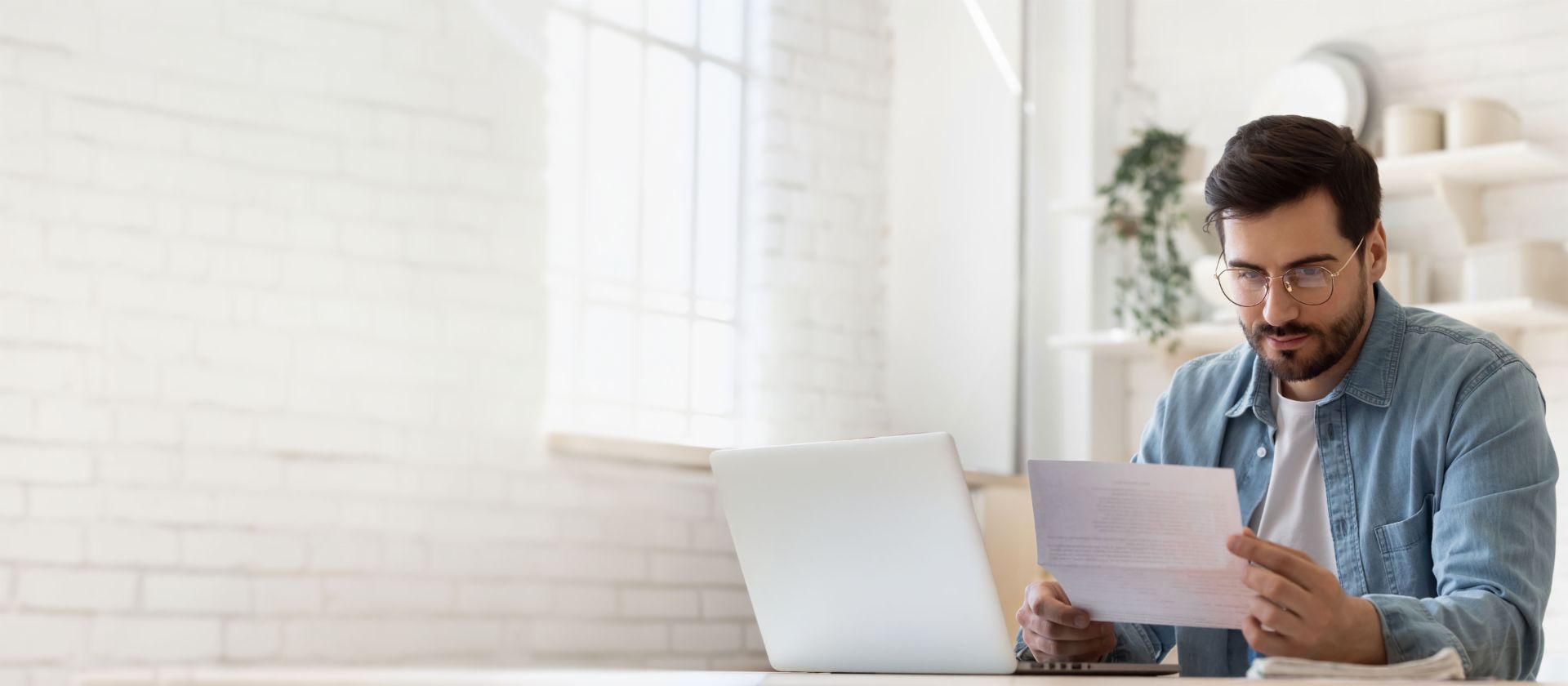 Bărbat cu ochelari lucrând la laptop în timp ce examinează documentul de reglementare a datoriilor într-o casă luminoasă
