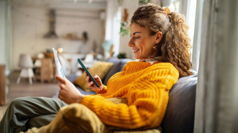 O tânără zâmbitoare cu păr creț, îmbrăcată într-un pulover galben confortabil, stă pe o canapea în sufrageria ei și se uită la smartphone-ul ei în timp ce ține un document de hârtie.