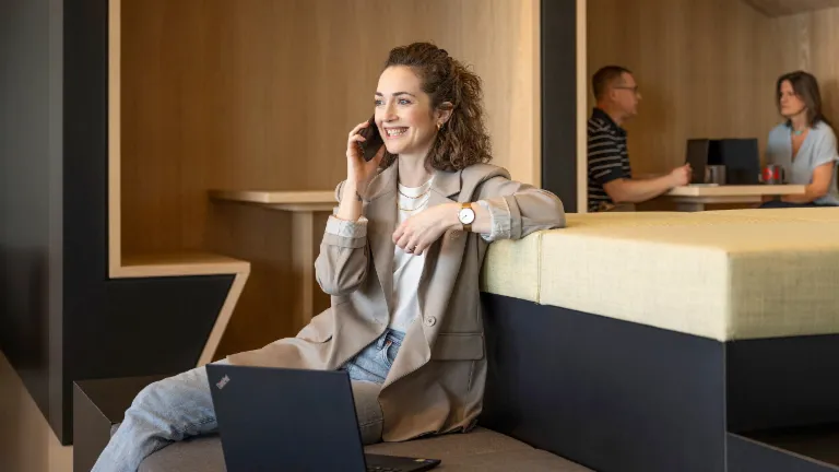 O persoană zâmbitoare stă într-un lounge modern de birou, vorbește la telefon și lucrează la un laptop. În fundal, alte două persoane lucrează.
