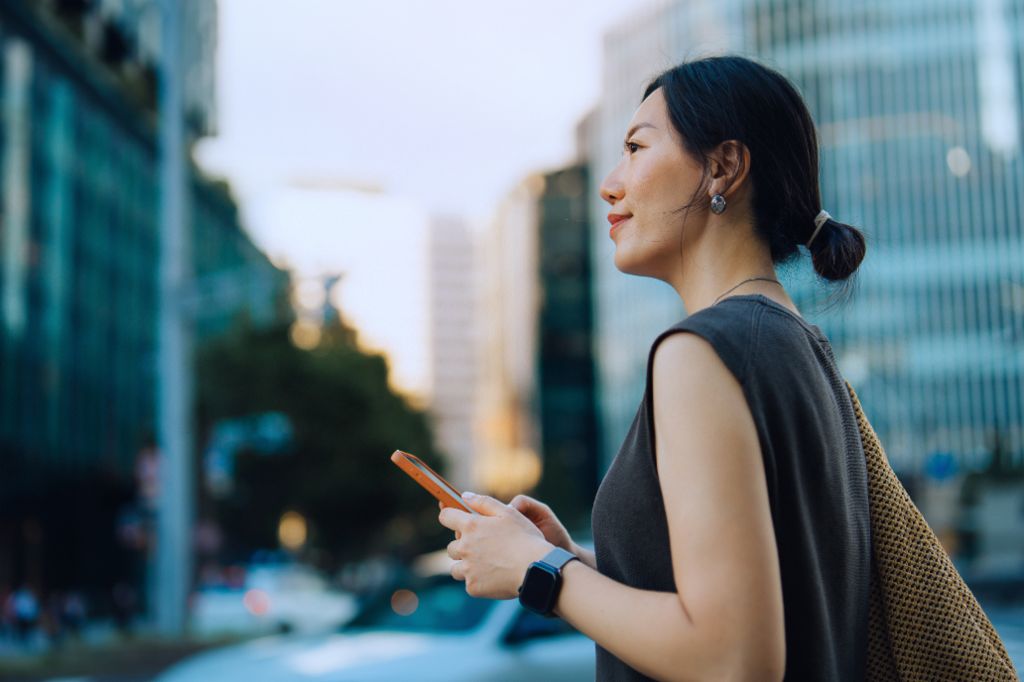 O tânără femeie stă în aer liber într-un mediu urban, ținând smartphone-ul și zâmbind în timp ce privește înainte. În fundal se văd clădiri moderne și copaci.