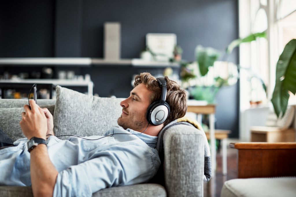 Un tânăr stă întins pe o canapea în sufrageria sa, poartă căști și se uită la smartphone-ul său zâmbind, bucurându-se de muzică sau de un podcast.