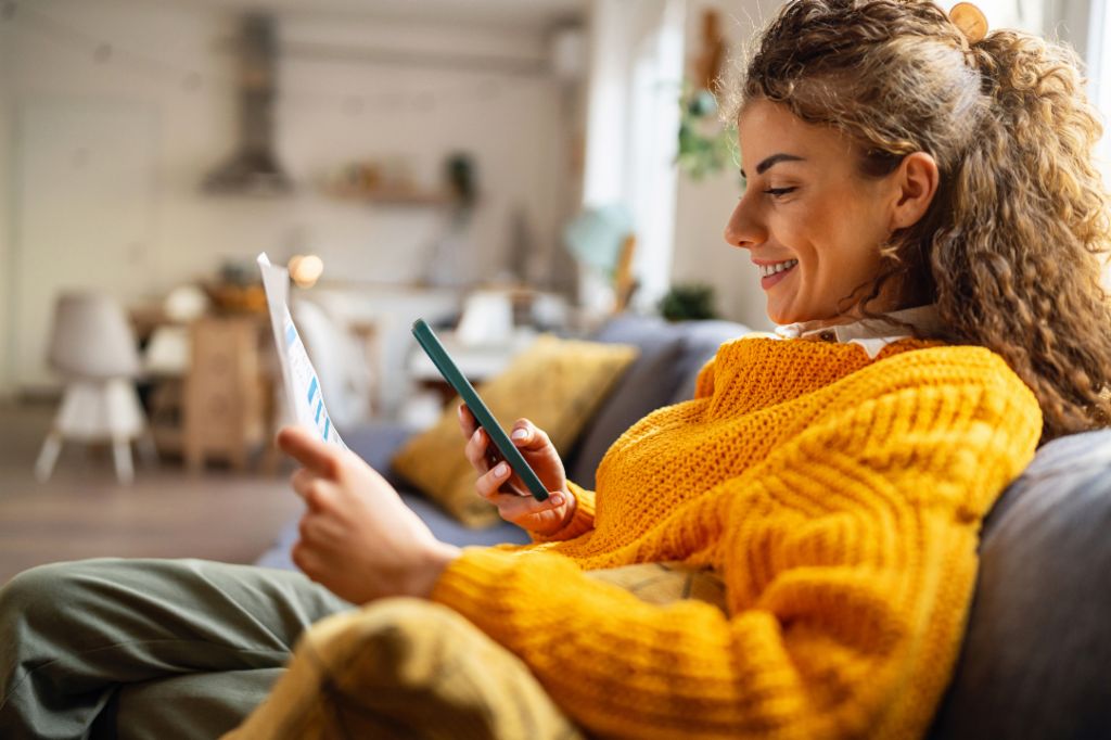 O tânără zâmbitoare cu păr creț, îmbrăcată într-un pulover galben confortabil, stă pe o canapea în sufrageria ei și se uită la smartphone-ul ei în timp ce ține un document de hârtie.