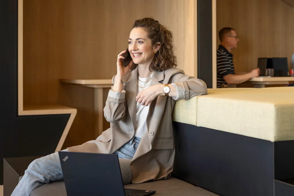 O persoană zâmbitoare stă într-un lounge modern de birou, vorbește la telefon și lucrează la un laptop. În fundal, alte două persoane lucrează.
