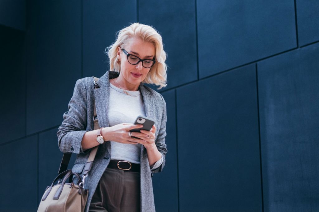 O femeie de afaceri blondă, cu blazer gri, stă în fața unei clădiri moderne și zâmbește uitându-se la smartphone-ul ei.