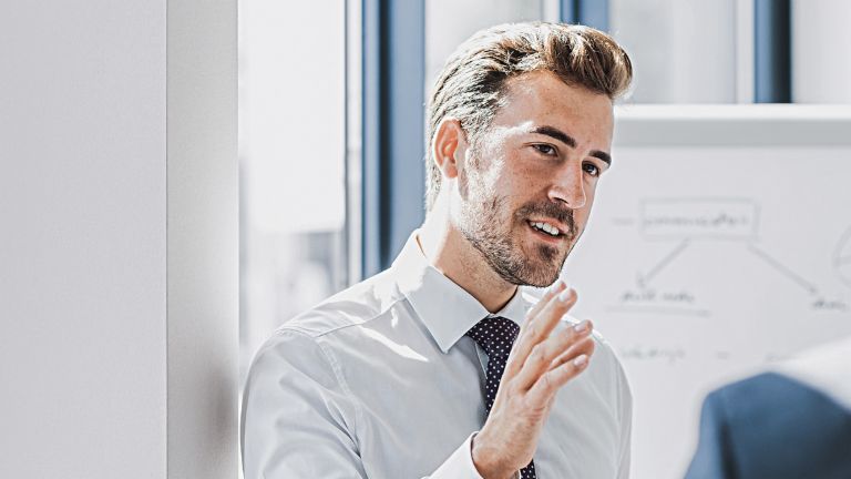 Se vede un bărbat în ținută business care explică ceva cu o expresie prietenoasă și implicată. În fața lui se află o altă persoană care îl ascultă cu atenție. În fundal se vede o tablă albă cu o schiță.