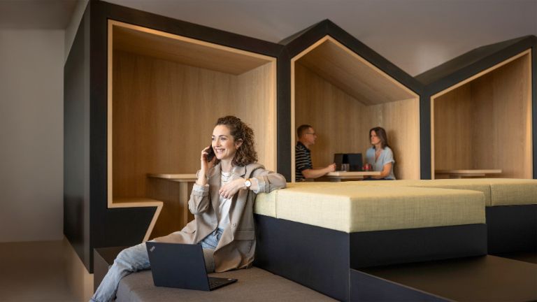 O persoană zâmbitoare stă într-un lounge modern de birou, vorbește la telefon și lucrează la un laptop. În fundal, alte două persoane lucrează.