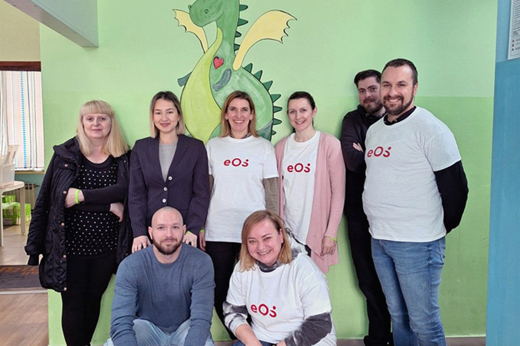 Opt persoane stau în fața unui perete pe care a fost pictat un dragon verde. Oamenii sunt aranjați în două rânduri: doi sunt ghemuiți în față și șase stau în picioare în spate. Patru persoane poartă tricouri albe cu sigla EOS roșie. Grupul zâmbește la aparatul foto. Pe podea, în față, pe dreapta, vedem câteva sticle și cutii de băuturi.