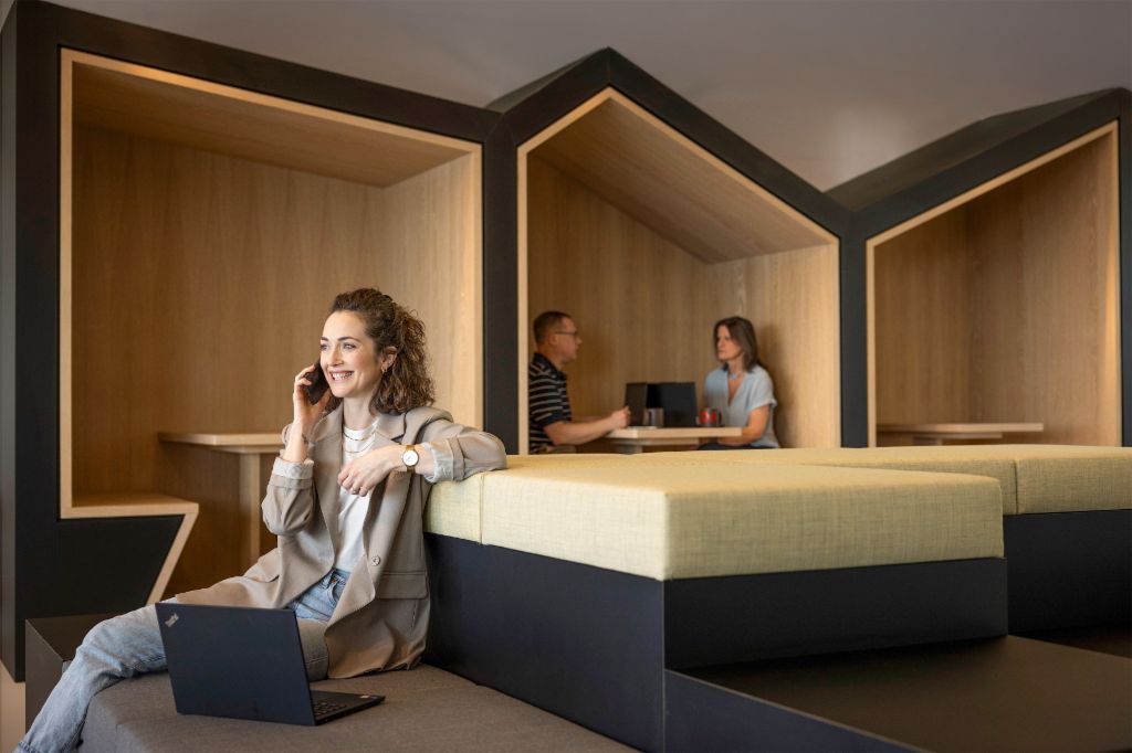 O persoană zâmbitoare stă într-un lounge modern de birou, vorbește la telefon și lucrează la un laptop. În fundal, alte două persoane lucrează.