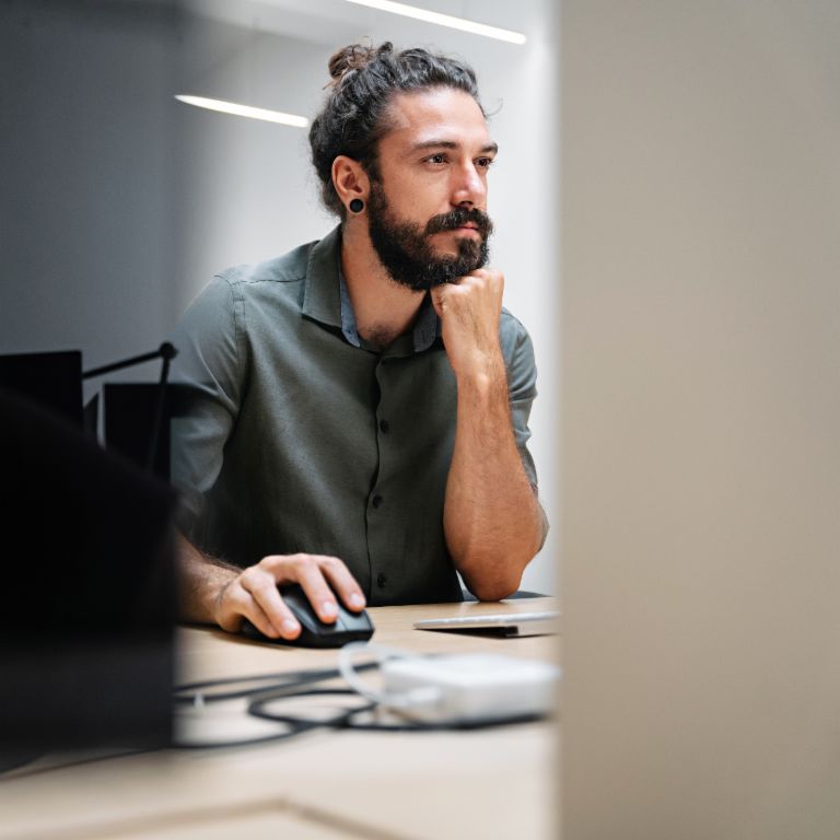 Un bărbat cu barbă și părul lung prins într-un coc stă la un computer, complet absorbit de munca sa.