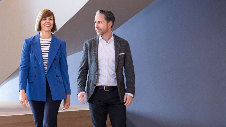 CEO-ul în blazer gri și CFO-ul Grupului EOS în blazer albastru trec printr-un birou modern. Spațiul dispune de feronerie mari, un perete curbat și lămpi suspendate elegante.