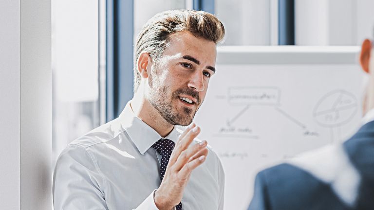 Se vede un bărbat în ținută business care explică ceva cu o expresie prietenoasă și implicată. În fața lui se află o altă persoană care îl ascultă cu atenție. În fundal se vede o tablă albă cu o schiță.