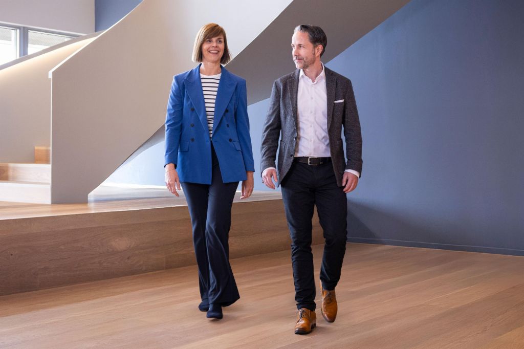 CEO-ul în blazer gri și CFO-ul Grupului EOS în blazer albastru trec printr-un birou modern. Spațiul dispune de feronerie mari, un perete curbat și lămpi suspendate elegante.